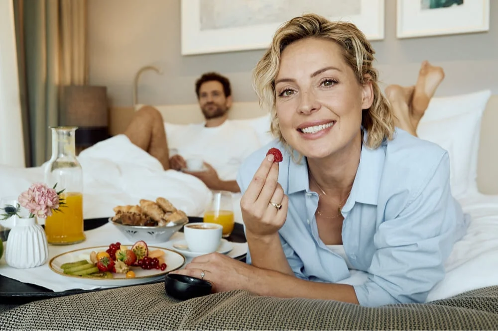 Frau liegt in Hotelbett und frühstückt Obst, ihr Mann liegt im Hintergrund Frau liegt in Hotelbett und frühstückt Obst, ihr Mann liegt im Hintergrund